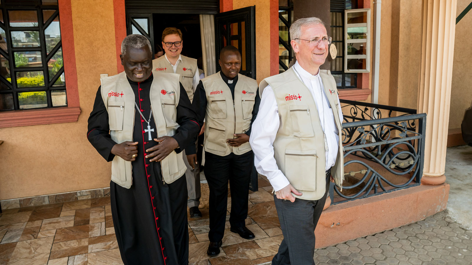 Erzbischof Philip Anyolo (links) und Erzbischof Dr. Stefan Heße (rechts) treten in Githurai im Großraum Nairobi aus einem Gebäude. Sie tragen beige Westen mit missio-Logo. Hinter ihnen folgen weitere Personen.