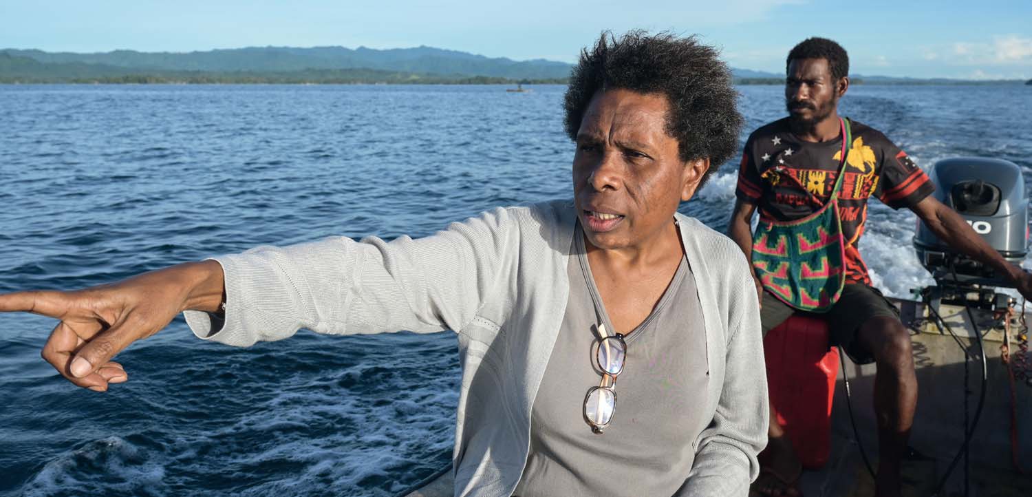 Rosa Koian sitzt in einem Boot und zeigt auf etwas vor ihr in der Ferne Liegendes. Ein Mann bedient den Außenbordmotor.