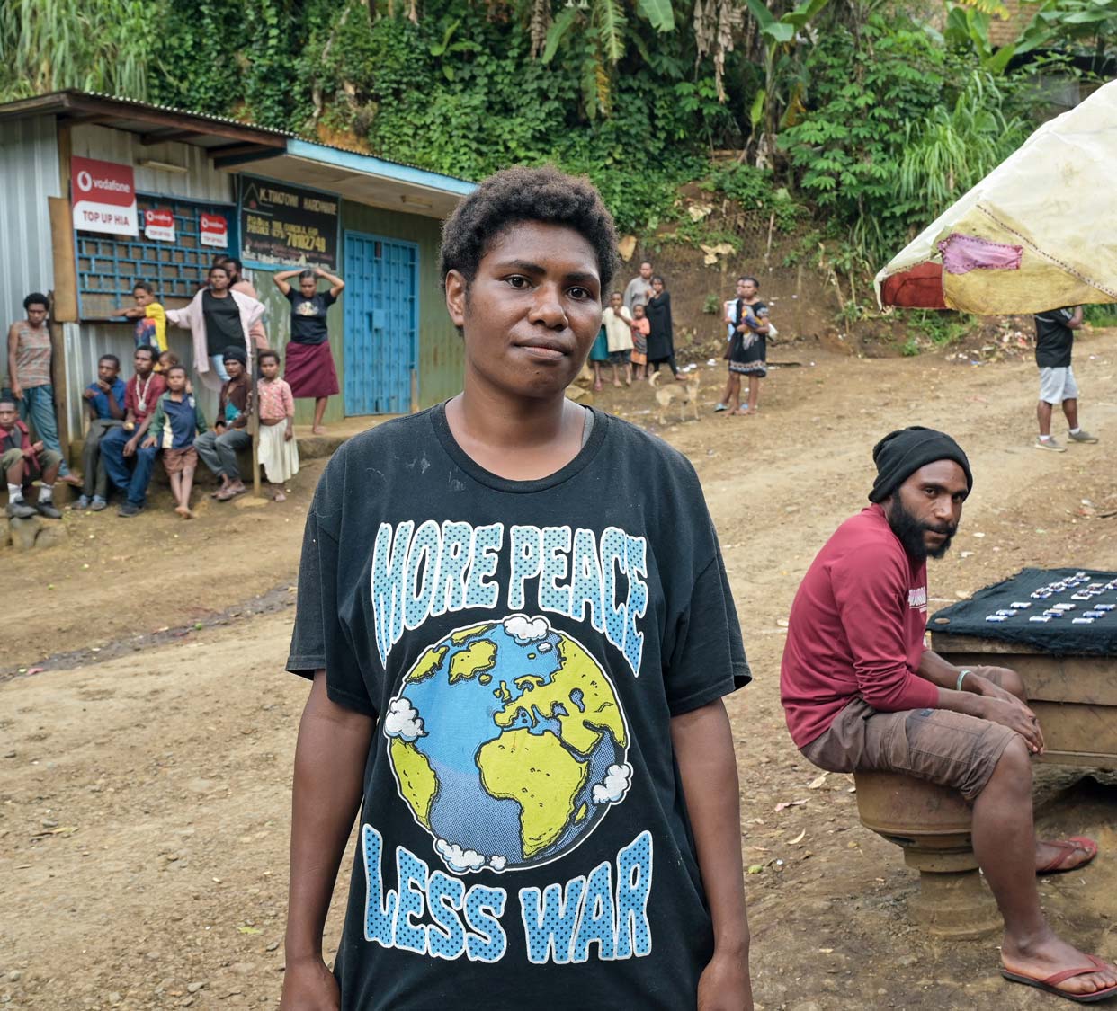 Im Vordergrund ein Jugendlicher mit einem T-Shirt, auf dem eine Weltkugel abgebildet ist und der Schriftzug Mehr Frieden, weniger Krieg steht. Im Hintergrund eine Straße, an deren Rand geschlossene Geschäfte und eine ganze Reihe von Leuten zu sehen sind.