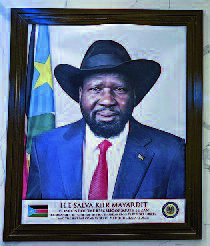 gerahmtes Bildnis des amtierenden Präsidenten Salva Kiir Mayardit. Er ist dargestellt in Anzug und Kravatte mit Hut, hinter ihm die Flagge des Südsudan.