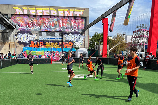 Spiel von zwei Streetfußball-Mannschaften auf einem auf dem Dach. Im Hintergrund eine Wand mit bunten Grafitti.