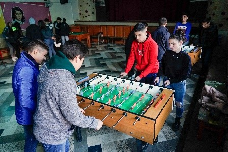 Eine Reihe von Jungs sowie ein paar Mädchen an zwei Tischkickern und zwei Billardtischen in einem Innenraum.