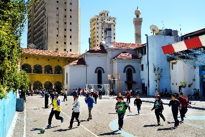 Kinder spielen Fußball auf einem Sportplatz im Hof. Hinter den Gebäuden, die den Innenhof umschließen, zeichnen sich vor dem blauen Himmel Hochhäuser ab.