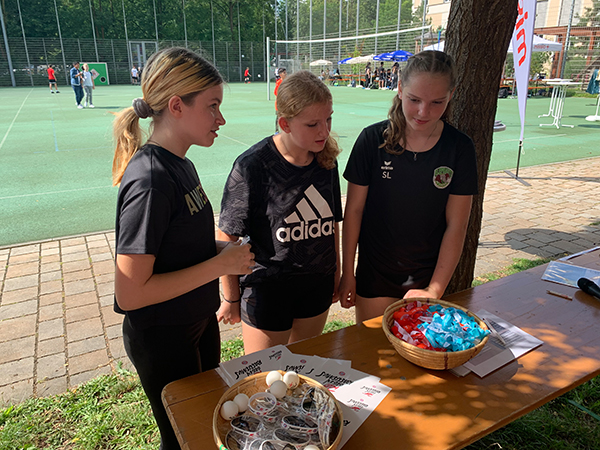 Drei Mädchen stehen am missio-Stand beim Unity-Cup. Auf dem Tisch vor ihnen liegen Armbänder mit Logo und Slogans von missio sowie vom FC Bayern und weitere Give-Aways.