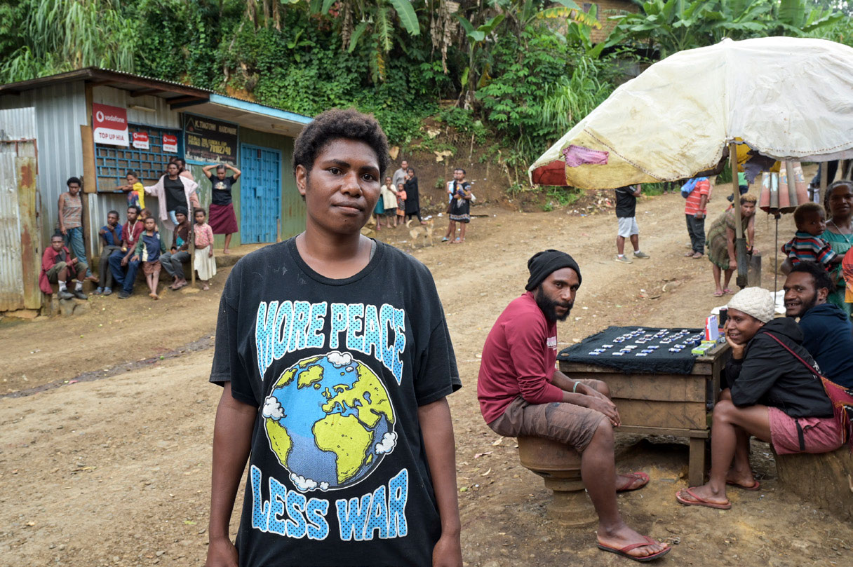 Straßenverkäufer im Settlement Pisswara, am Stadtrand von Goroka. Im Vordergrund auf ungeteerter Straße steht eine Frau und sieht skeptisch in die Kamera.