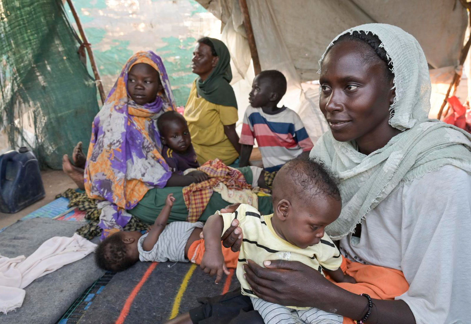 Babajenana sitzt mit einem Kind auf dem Schoß zusammen mit weiteren Geflüchteten in einem provorisorischen Zelt im Flüchtlingscamp im Südsudan.