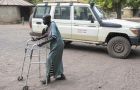 Eine Frau ist mit Gehwagen vor einem Geländewagen der Diözese Wau zu sehen.