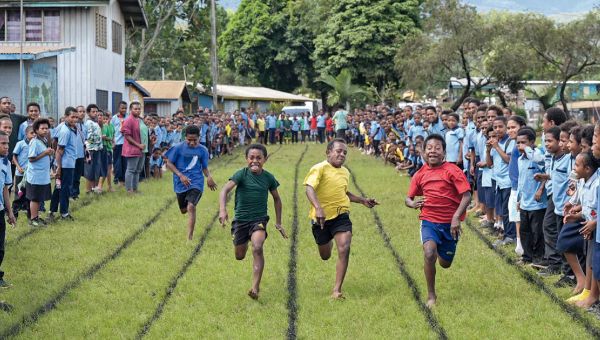 Vier Jungs sprinten um die Wette. Die Laufbahnen sind auf die Wiese gesprüht. Rechts und links davon feuern eine Vielzahl von Kindern und Jugendlichen in Schuluniform die Läufer an.